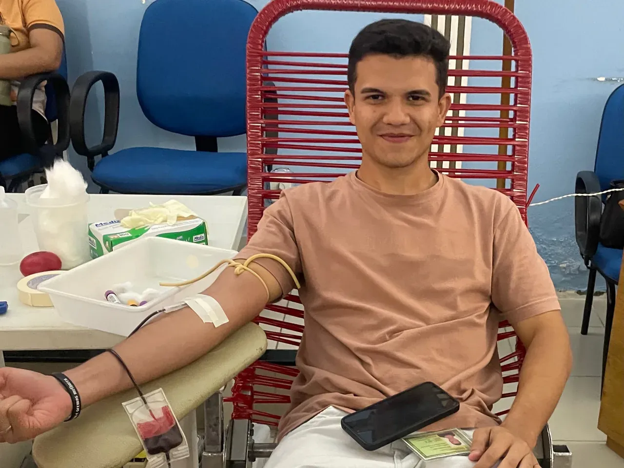 Jovem sentado na poltrona de doação sorri com o braço na maca, durante uma coleta de sangue